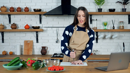 Crazy housewife in beige apron playing knife game on cutting board. Beautiful woman stands at home near kitchen table with ingredients vegetables for delicious healthy salad and laptop computer