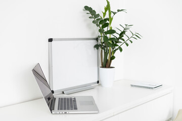 Close up view of simple workspace with laptop, notebooks and tree pot on white table with blurred office room background