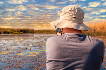 Young photographer man with a hat rides a traditional mokoro boat on the Okavango delta and takes picture at sunset. Botswana