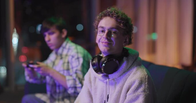 Attractive curly-haired caucasian young adult wearing headphones smiling cheerful into camera staying with best friend in living room playing games spending freetime.