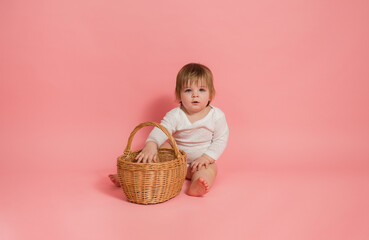 little girl in a white bodysuit sits with a wicker basket on a pink background with space for text