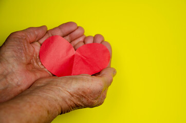 Fototapeta premium Heart in old palms. Heart in old wrinkled hands. Heart on a yellow background. Love concept. Caring and kindness. Charity. Grandmother with a heart in her hands. Mothers Day. Women's Day