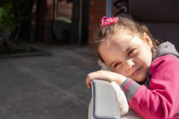 A pensive serious preschool girl looks to the side, a pensive child sits on the street, holds his hands on his cheek, dreams, asks questions, a child's profile in the fresh air.