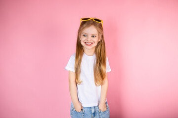 Little girl in sunglasses with long hair standing on a pink background with her hands in her pockets