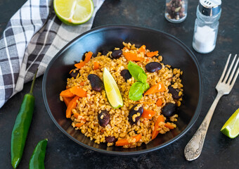 Bulgur pilaf with carrots and black beans in a black plate on a dark concrete background. Vegetarian recipes.