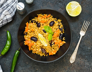 Bulgur pilaf with carrots and black beans in a black plate on a dark concrete background. Vegetarian recipes.