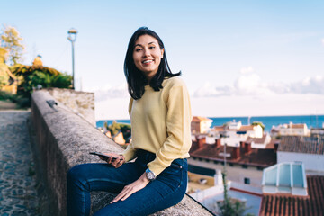 Fototapeta na wymiar Portrait of cheerful Asian woman dressed in casual clothing smiling at camera during travel vacations, happy female tourist with modern cellphone device in hand posing at overlooking street area