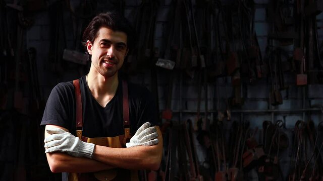 Authentic blacksmith man forges a metal product looking at the camera and arms crossed in dark indoors studio feel confidence