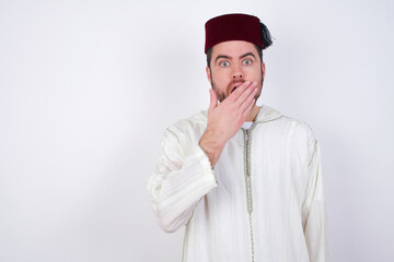 Emotional young handsome Caucasian man wearing Arab djellaba and Fez hat over white wall gasps from astonishment, covers opened mouth with palm, looks shocked at camera. Reaction concept