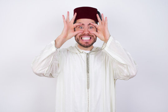 young handsome Caucasian man wearing Arab djellaba and Fez hat over white wall keeping eyes opened to find a success opportunity.