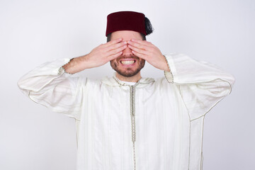 young handsome Caucasian man wearing Arab djellaba and Fez hat over white wall covering eyes with...