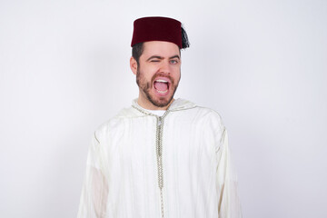young handsome Caucasian man wearing Arab djellaba and Fez hat over white wall winking looking at...