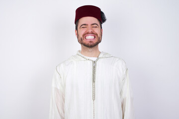 young handsome Caucasian man wearing Arab djellaba and Fez hat over white wall very happy and excited about new plans.