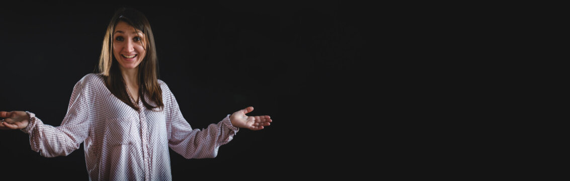 portrait of young surprised woman isolated on black wide background. Image of caucasian brunette woman wearing casual t-shirt shrugging and throwing up her hands isolated