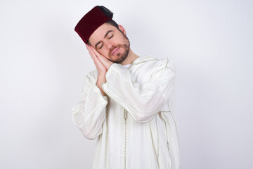 young handsome Caucasian man wearing Arab djellaba and Fez hat over white wall sleeping tired dreaming and posing with hands together while smiling with closed eyes.