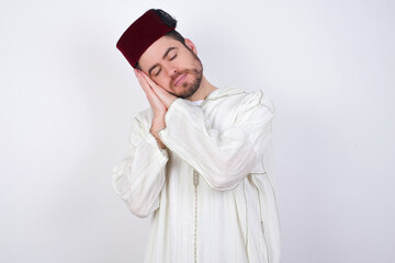 Fototapeta premium young handsome Caucasian man wearing Arab djellaba and Fez hat over white wall sleeping tired dreaming and posing with hands together while smiling with closed eyes.
