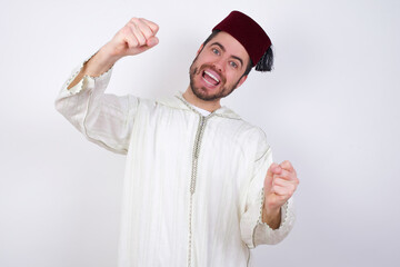 Naklejka premium young handsome Caucasian man wearing Arab djellaba and Fez hat over white wall imagine steering wheel helm rudder passing driving exam good mood fast speed