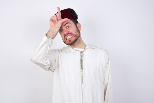 young handsome Caucasian man wearing Arab djellaba and Fez hat over white wall making fun of people with fingers on forehead doing loser gesture mocking and insulting.