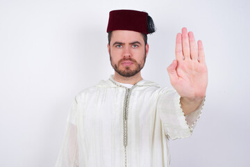 young handsome Caucasian man wearing Arab djellaba and Fez hat over white wall doing stop sing with palm of the hand. Warning expression with negative and serious gesture on the face.