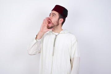 young handsome Caucasian man wearing Arab djellaba and Fez hat over white wall shouting and screaming loud to side with hand on mouth. Communication concept.