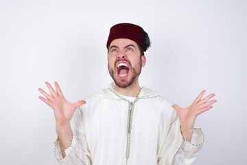 young handsome Caucasian man wearing Arab djellaba and Fez hat over white wall crazy and mad shouting and yelling with aggressive expression and arms raised. Frustration concept.