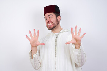 young handsome Caucasian man wearing Arab djellaba and Fez hat over white wall afraid and terrified with fear expression stop gesture with hands, shouting in shock. Panic concept.