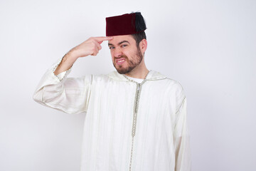 young handsome Caucasian man wearing Arab djellaba and Fez hat over white wall pointing unhappy at...