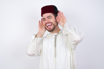 young handsome Caucasian man wearing Arab djellaba and Fez hat over white wall Trying to hear both hands on ear gesture, curious for gossip. Hearing problem, deaf