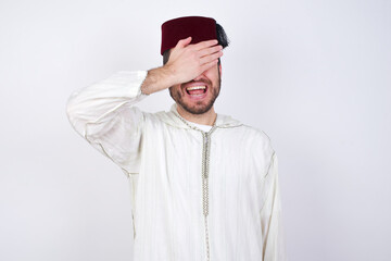 young handsome Caucasian man wearing Arab djellaba and Fez hat over white wall smiling and laughing with hand on face covering eyes for surprise. Blind concept.