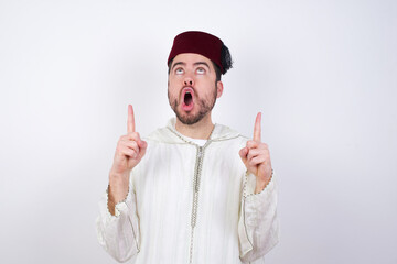 young handsome Caucasian man wearing Arab djellaba and Fez hat over white wall amazed and surprised...