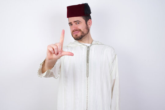 young handsome Caucasian man wearing Arab djellaba and Fez hat over white wall making fun of people with fingers on forehead doing loser gesture mocking and insulting.