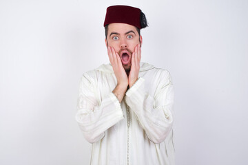 Scared terrified young handsome Caucasian man wearing Arab djellaba and Fez hat over white wall shocked with prices at shop, People and human emotions concept