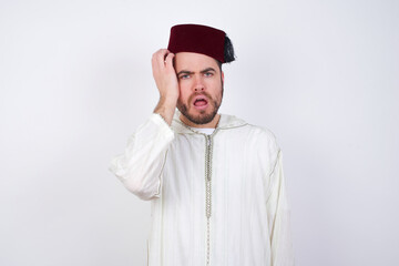 Fototapeta premium Embarrassed young handsome Caucasian man wearing Arab djellaba and Fez hat over white wall with shocked expression, expresses great amazement, Puzzled model poses indoor