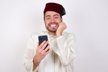 Joyous young handsome Caucasian man wearing Arab djellab poses with mobile phone device, types text message on modern smartphone, watches funny video during free time, enjoys good internet connection,