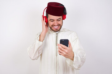 Happy young handsome Caucasian man wearing Arab djellaba and Fez hat over white wall feels good while focused in screen of smartphone. People, technology, lifestyle