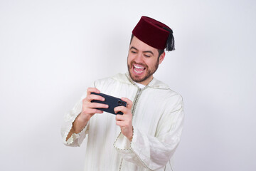 young handsome Caucasian man wearing Arab djellaba and Fez hat over white wall holding in hands cell playing video games or chatting