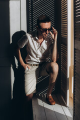 Stylish guy, model, in stylish casual shorts and a shirt in the studio with glasses, summer fashion