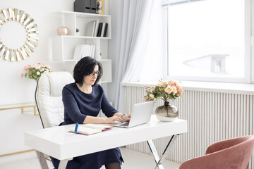 Fototapeta na wymiar a young woman in glasses and in a blue shirt works at home at a laptop as a coach in front of the kitchen in a bright interior, a concept of a workplace and remote work at home