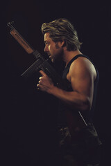 Rugged man with machine gun in dark blue tank top against dark background.