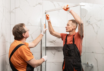 Workers are connecting the glass walls of the shower enclosure with a metal bar.