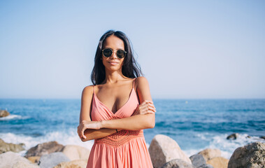 Fototapeta na wymiar Half length portrait of carefree female tourist in sundress and glasses for sun protection looking at camera while posing at coastline, charming woman in casual wear spending day for promenade