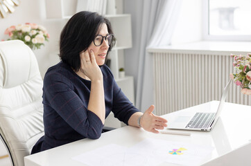 Obraz na płótnie Canvas a young woman in glasses and in a blue dress works at home at a laptop as a coach in a bright interior, a concept of a workplace and remote work at home