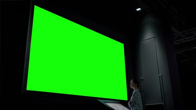Mock up, futuristic, chroma key, template, green screen concept. Woman using electronic kiosk and looking at green blank large interactive wall display in dark room of modern technology museum