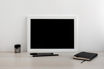 White frame with black background in a bright room on the desktop. Notebook and pens in the workplace. Minimalistic design