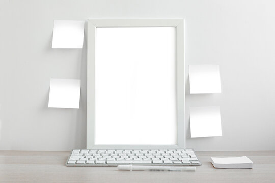 White picture frame in a bright room on the desktop. Notebook, self-adhesive notes, computer keyboard and pens in the workplace. Minimalistic design
