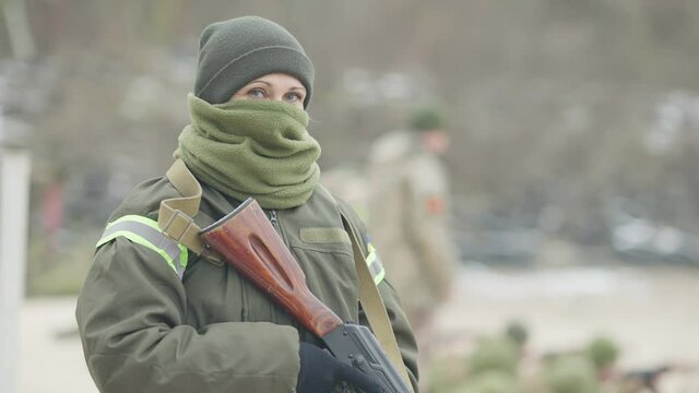Portrait of confident masked military woman standing on polygon holding rifled gun. Beautiful Caucasian woman looking at camera and turning away. Army and equality concept.