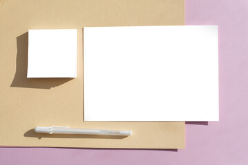 A blank layout of a white sheet on an abstract pink and beige table illuminated by the sun. The concept of minimalism and modernity. White pen under the sheet and paper for notes.