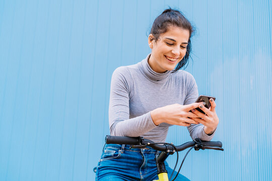 Cheerful beautiful woman using mobile phone while sitting on fixie bike against blue wall