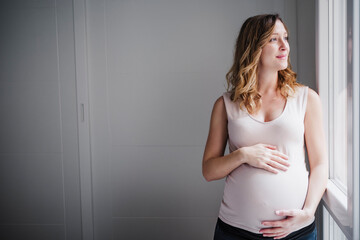 Thoughtful pregnant woman with hands on stomach looking through window at home