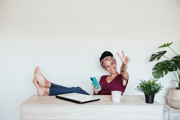 Cheerful female entrepreneur making peace sign while relaxing with feet up at home office